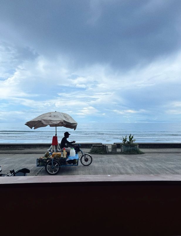 "A food merchant on a Tuktuk, somewhere at the sea side. His vehicle is protected from the sun with a parasol."